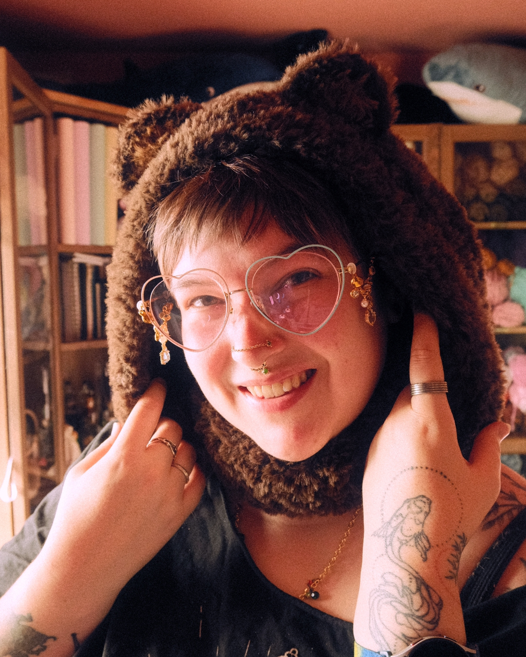 Person wearing a brown bear-themed hat with ears, heart-shaped glasses, and multiple rings in a cozy room.