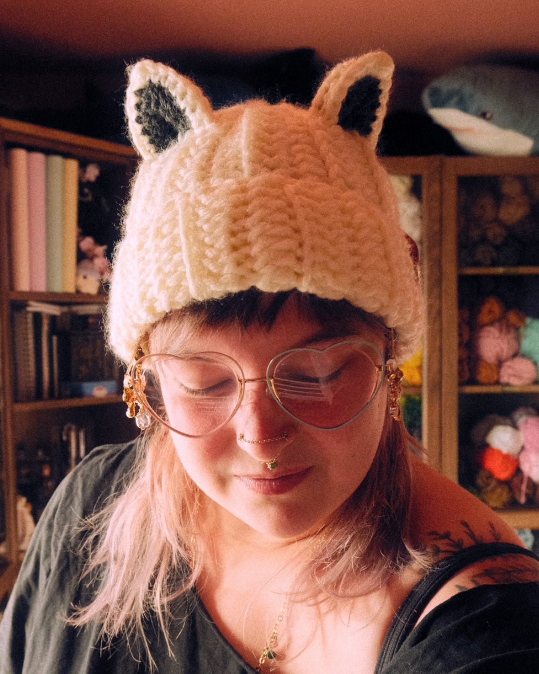 Person wearing a crochet hat with arctic fox ears in a cozy room with bookshelves and stuffed animals.