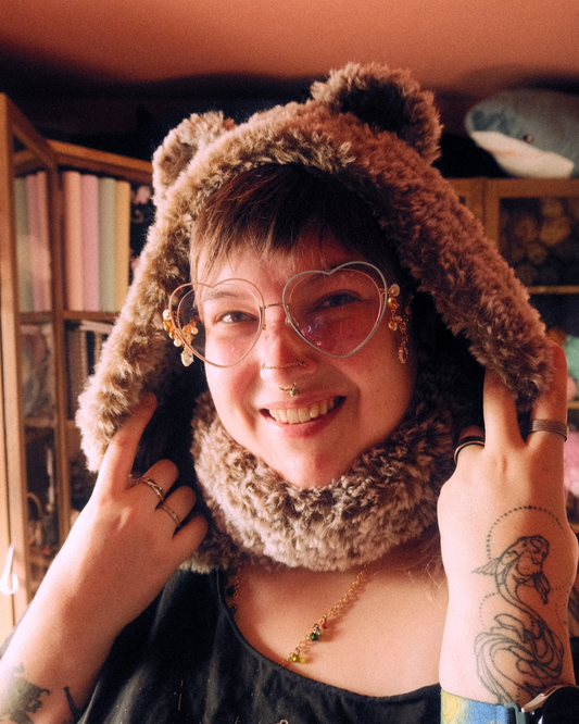 Person wearing a brown crochet hood with bear ears and heart-shaped glasses, smiling.