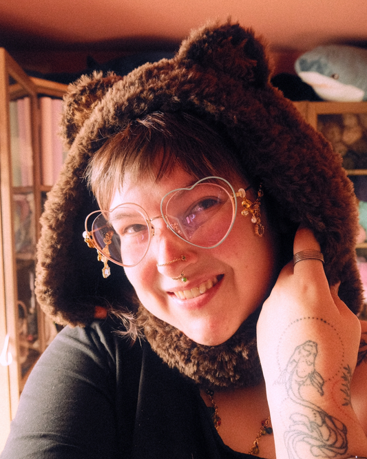 Person wearing a brown hood with bear ears, heart-shaped glasses, and a tattoo on their arm.