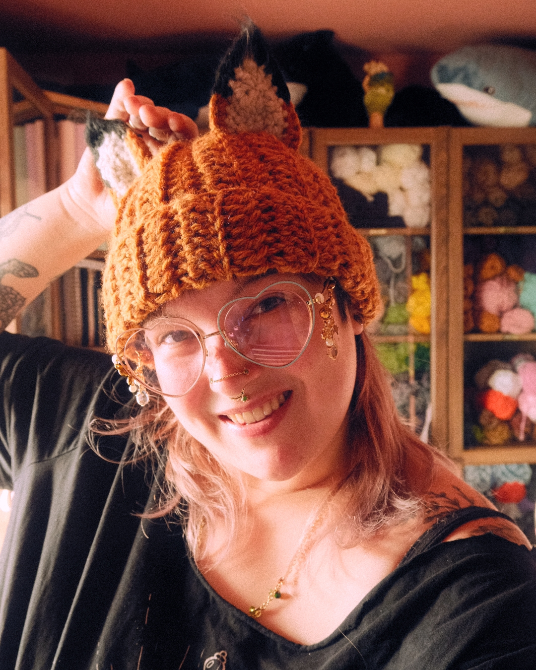 Person wearing a crochet fox hat with ears in a cozy room filled with plush toys.