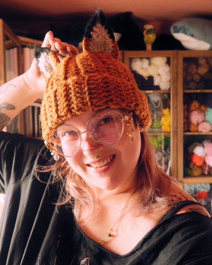 Person wearing a crochet fox hat with ears in a cozy room filled with plush toys.