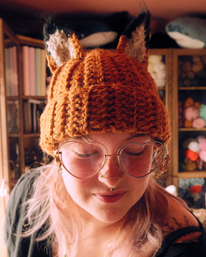 Person wearing a crochet fox hat in a cozy room with shelves in the background
