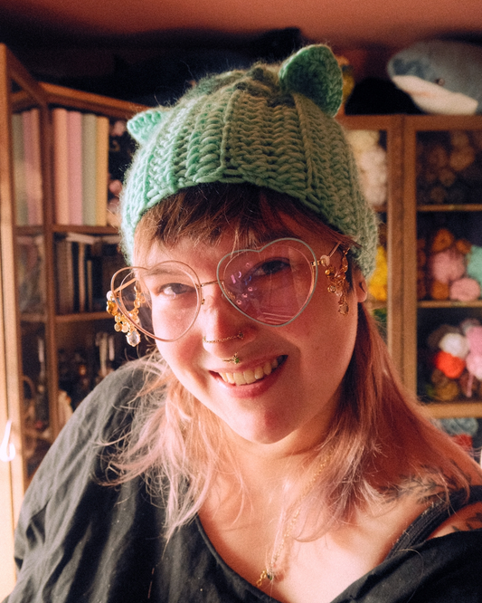 Person wearing a green knitted hat with gently pointed ears in a room with shelves and toys.