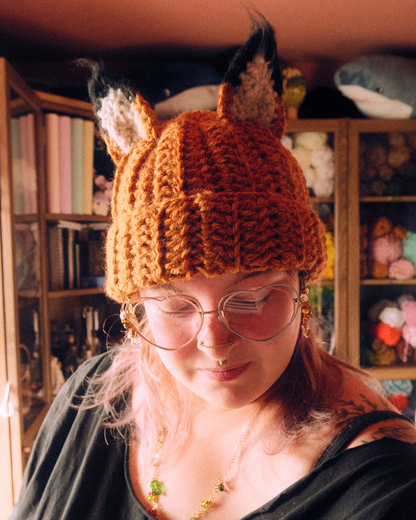 Person wearing a crochet fox hat in a cozy room with bookshelves and toys.